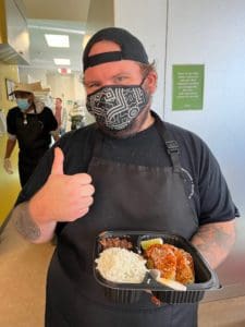 Masked chef posing for photo while holding food tray and giving thumbs up